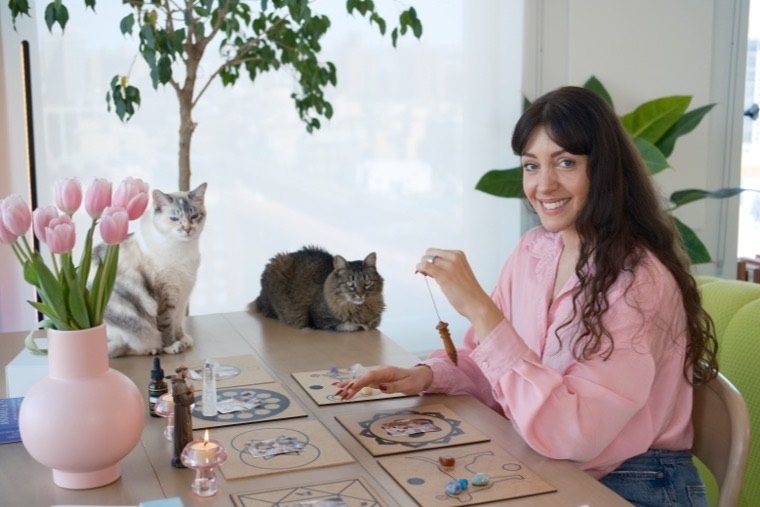 Bruna at her table by the window, smiling, with her two cats beside her and the chakra boards laid out